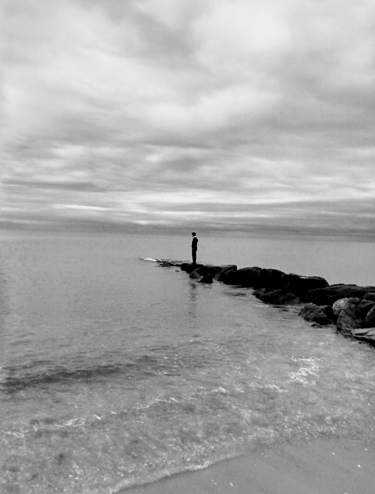 black and white photo of a person standing on sea rocks on a cloudy day