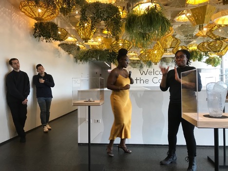 Curator Adero Knott, resplendent with her golden dress beneath the hanging plants, introduces the exhibits, including the translucent prosthesis visible in this picture. The ASL interpreter signs to a deaf audience member. Another exhibit shows the purple mold for a hearing aid.