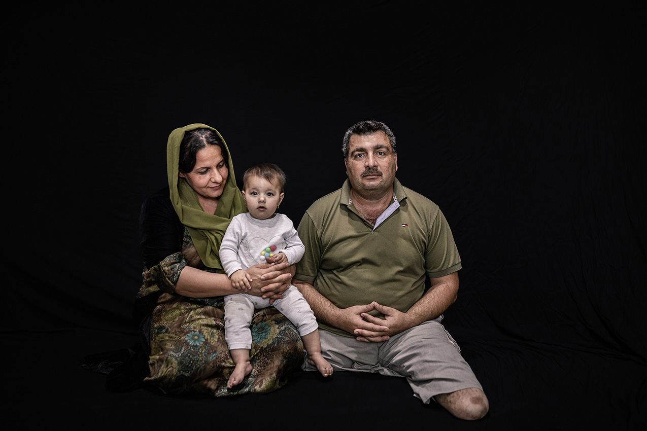 Portrait of a Kurdish family (man, woman and baby) on a black background. The man has both legs amputated.