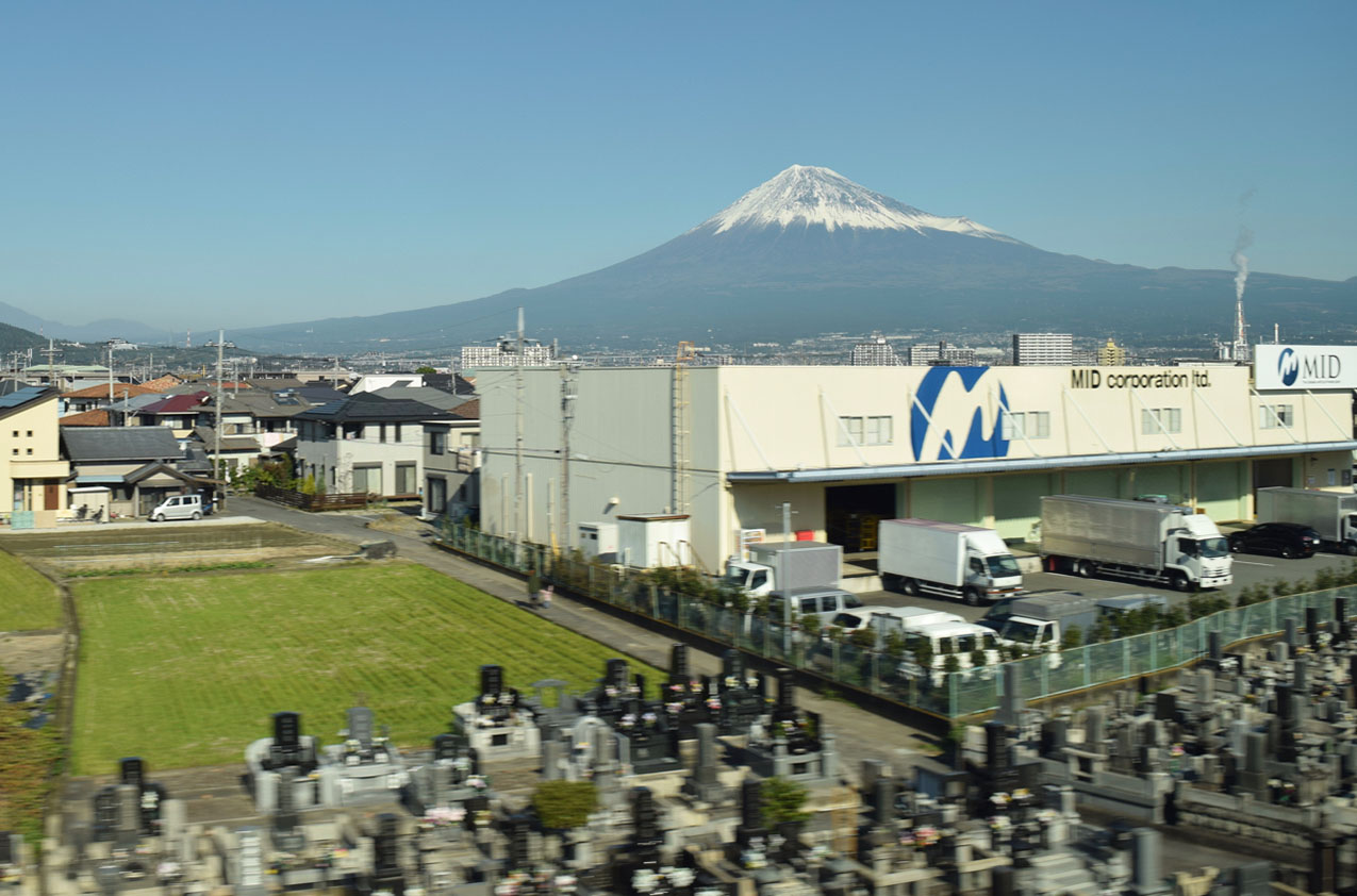 Mount Fuji in the distance with a cemetary in the foreground and industrial and residential buildings in the midground