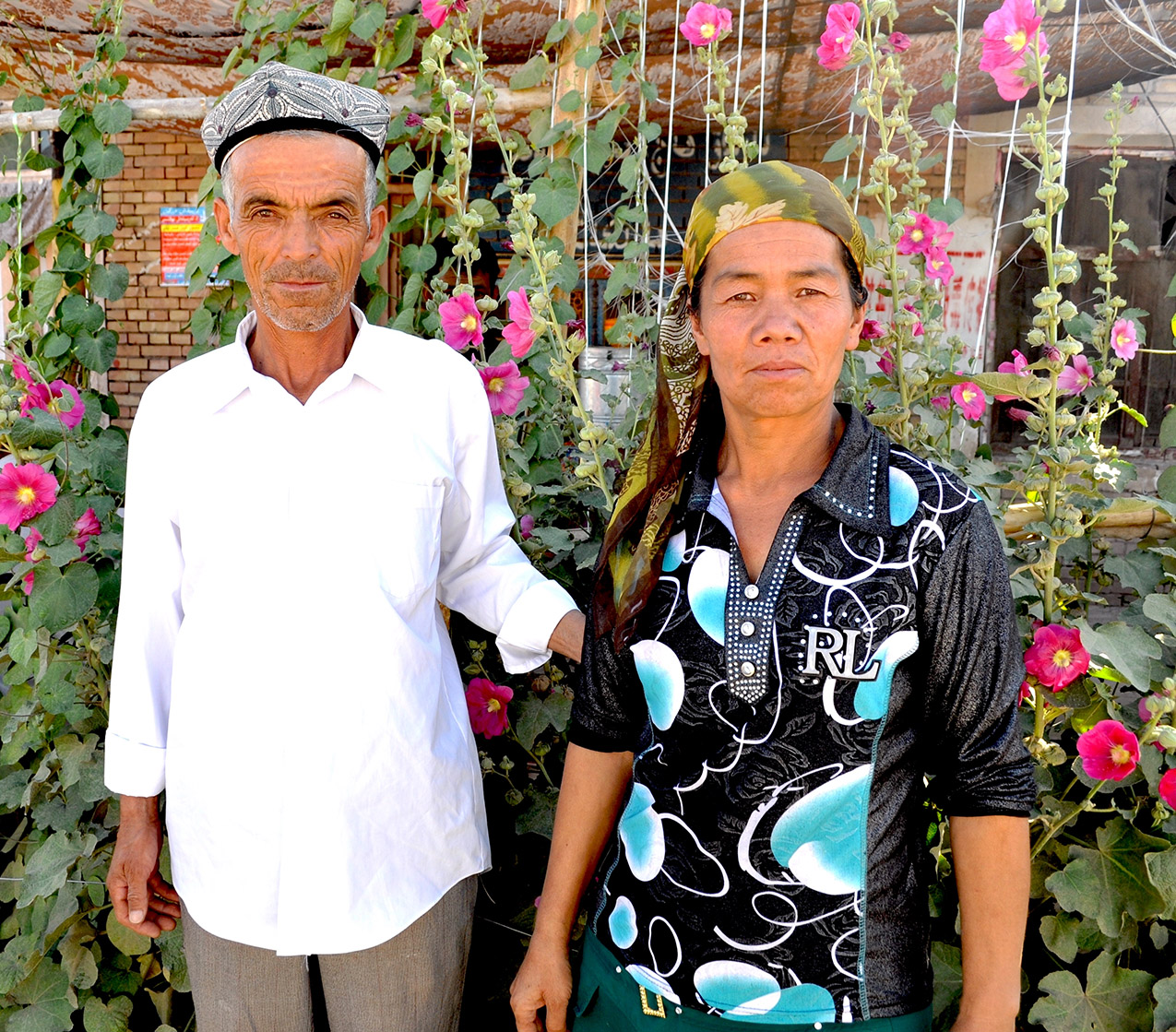 A man in a white shirt and a woman with a headscarf wearing a Ralph Lauren shirt in front of a trellis of pink hollyhocks in Kashgar, China