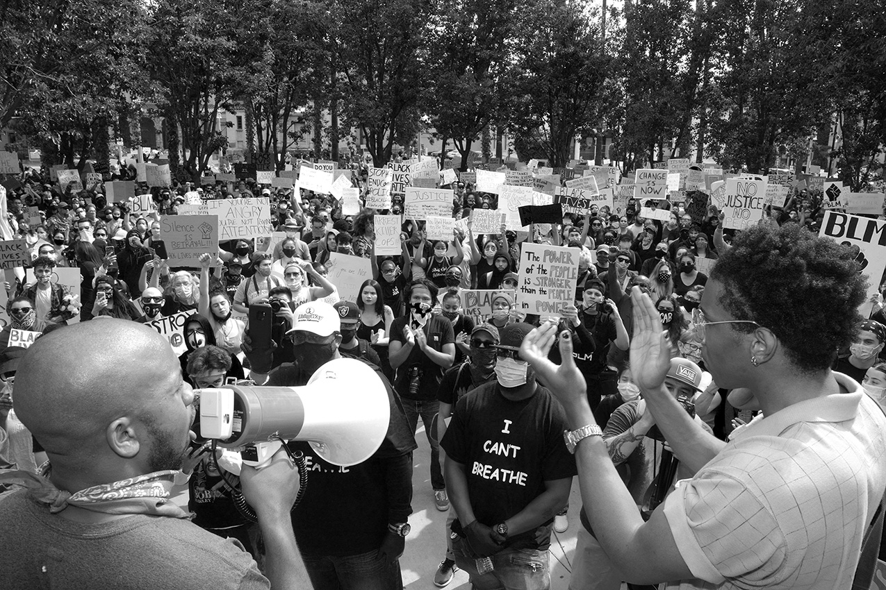 a Black man on a megaphone addresses a crowd of clapping protesters holding signs demanding racial justice