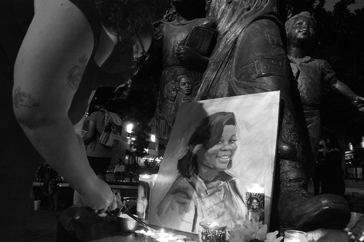 black and white photo of a woman lighting tealight candles at a makeshift altar featuring a painting of Breonna Taylor