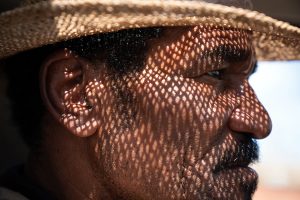 Closeup profile of a middle-aged Latino man wearing a straw hat; the sun casts diamond-shaped shadows onto his face