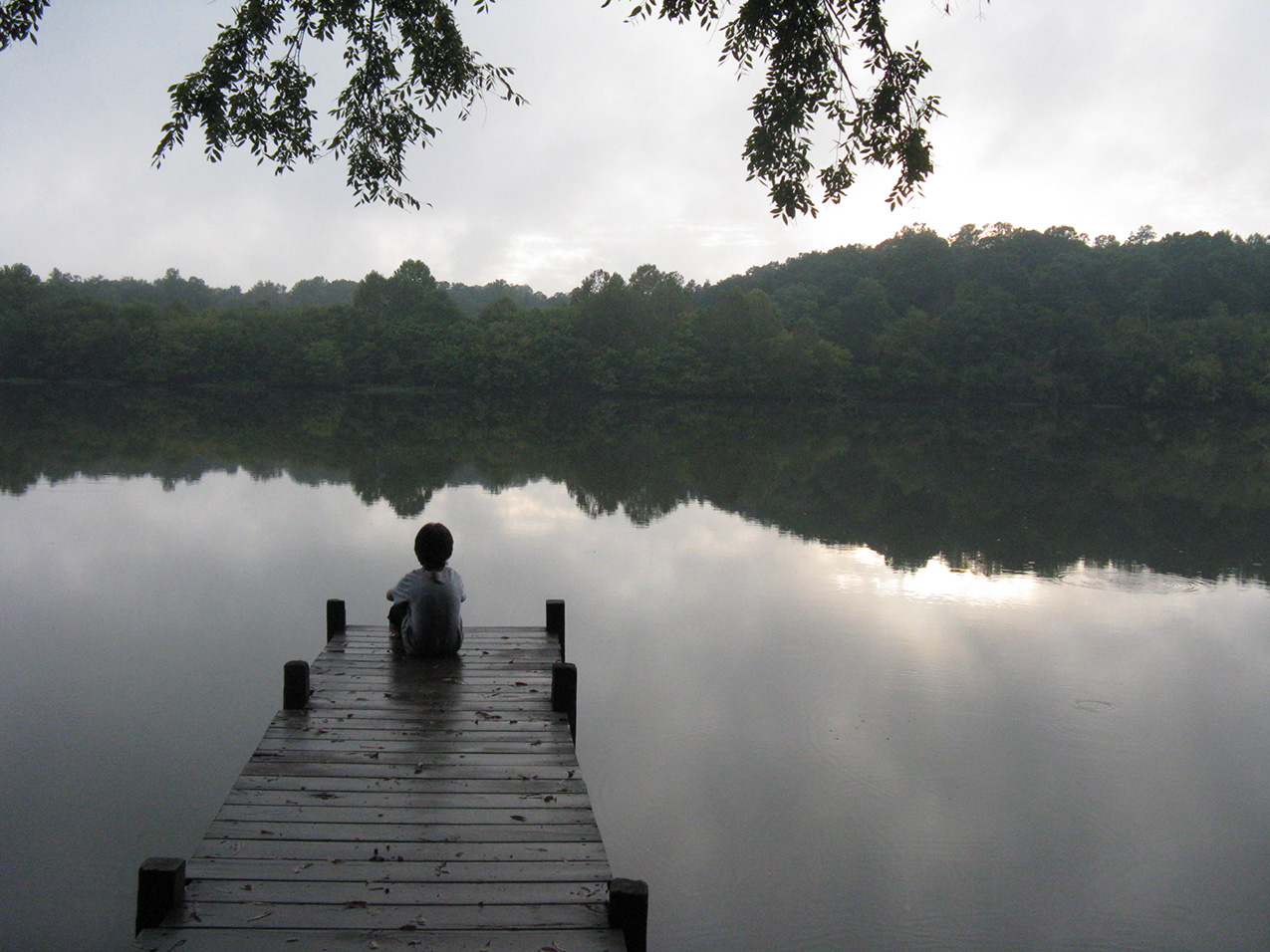 a small boy, seen from behind, sits on a dock with his knees hugged to his chest. the river is still, the sky is gray and cloudy.