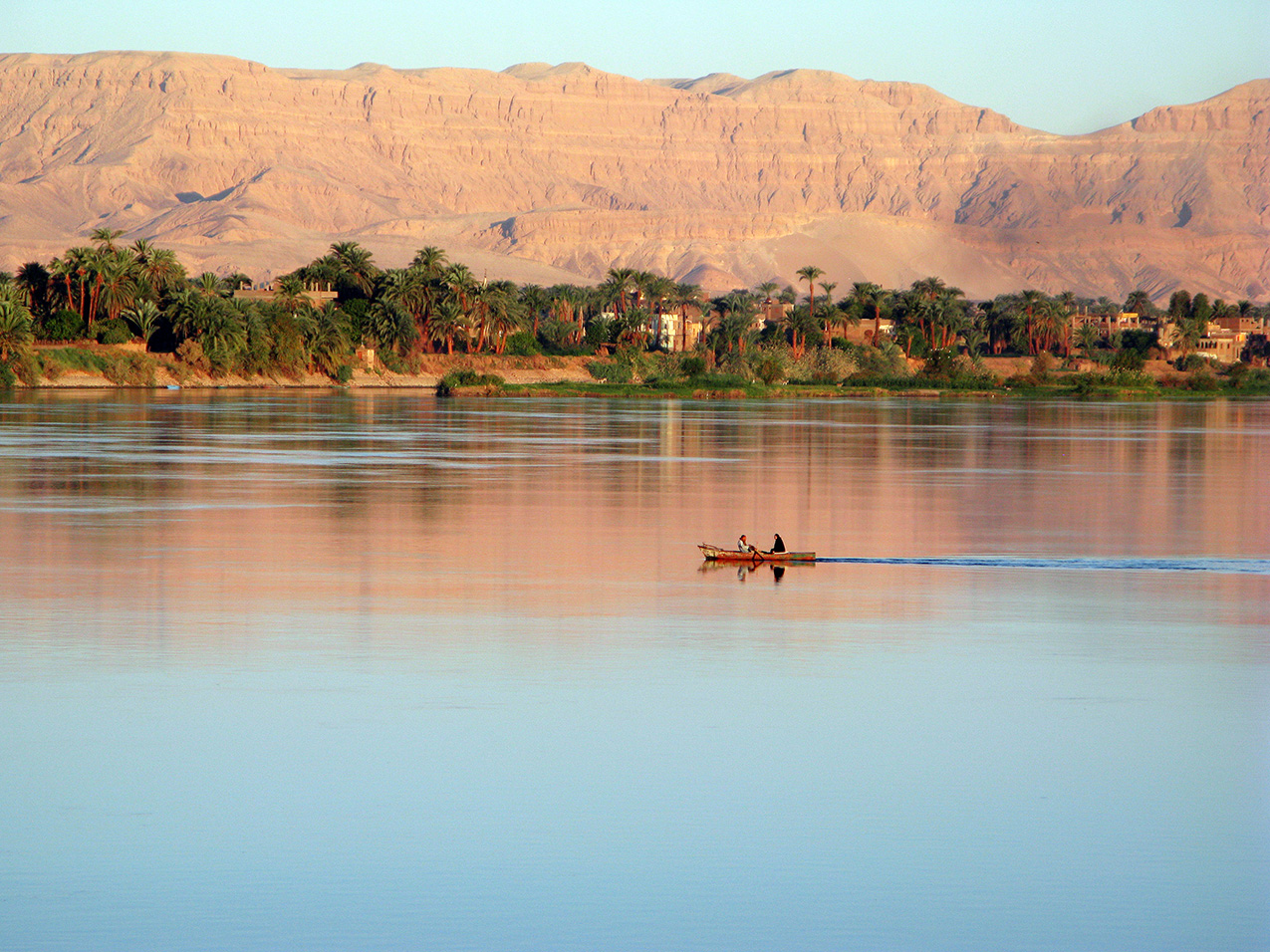 two people travel in a red boat on the Nile River in Egypt. the water is blue but reflects the orange cliffs in the distance, and palm trees and buildings cover the opposite shore.