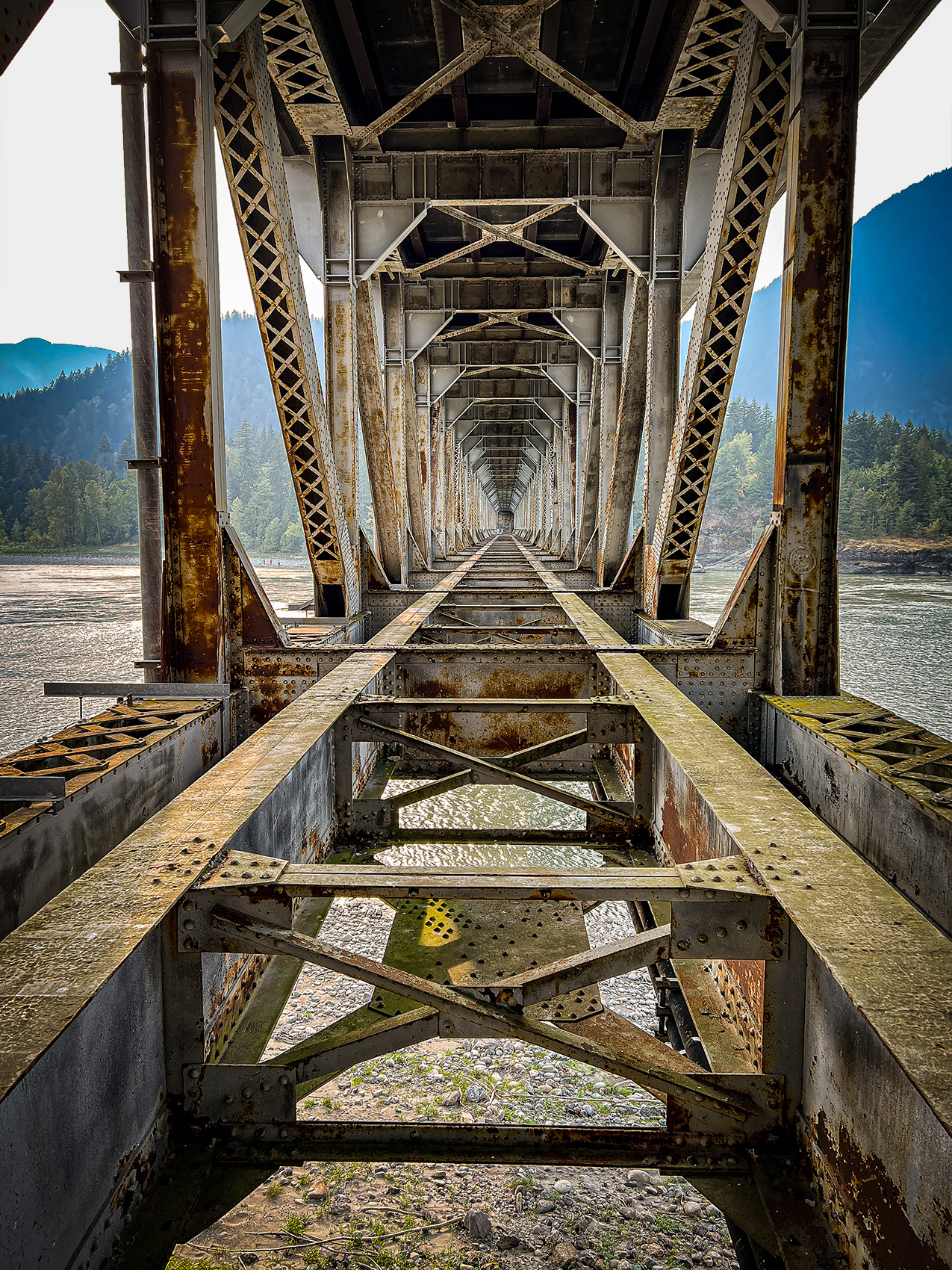 symmetrical, one-point perspective photo of the underside of a gritty metal bridge over the Fraser River at Hope, British Columbia