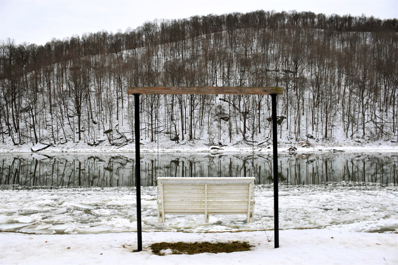 a white bench swing in front of a river. the ground is covered in snow, the trees are bare and reflect in the still water.