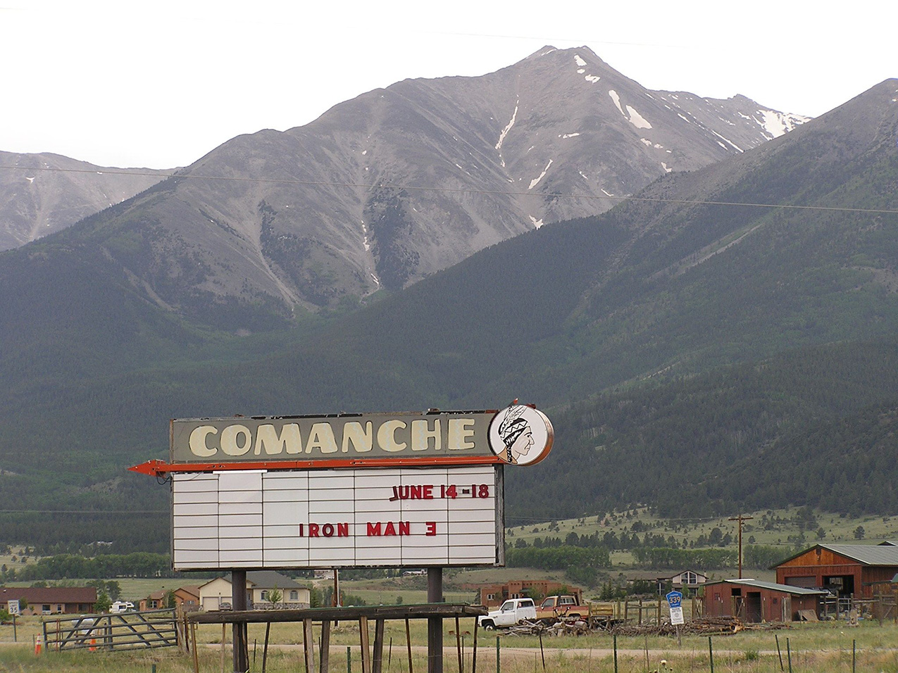 a vintage marquee sign with COMANCHE and a cartoon of a Native American man at the top displays IRON MAN 3 JUNE 14-18; behind the sign are scattered houses and junked pickup trucks dwarfed by mountains that rise suddenly from the foothills
