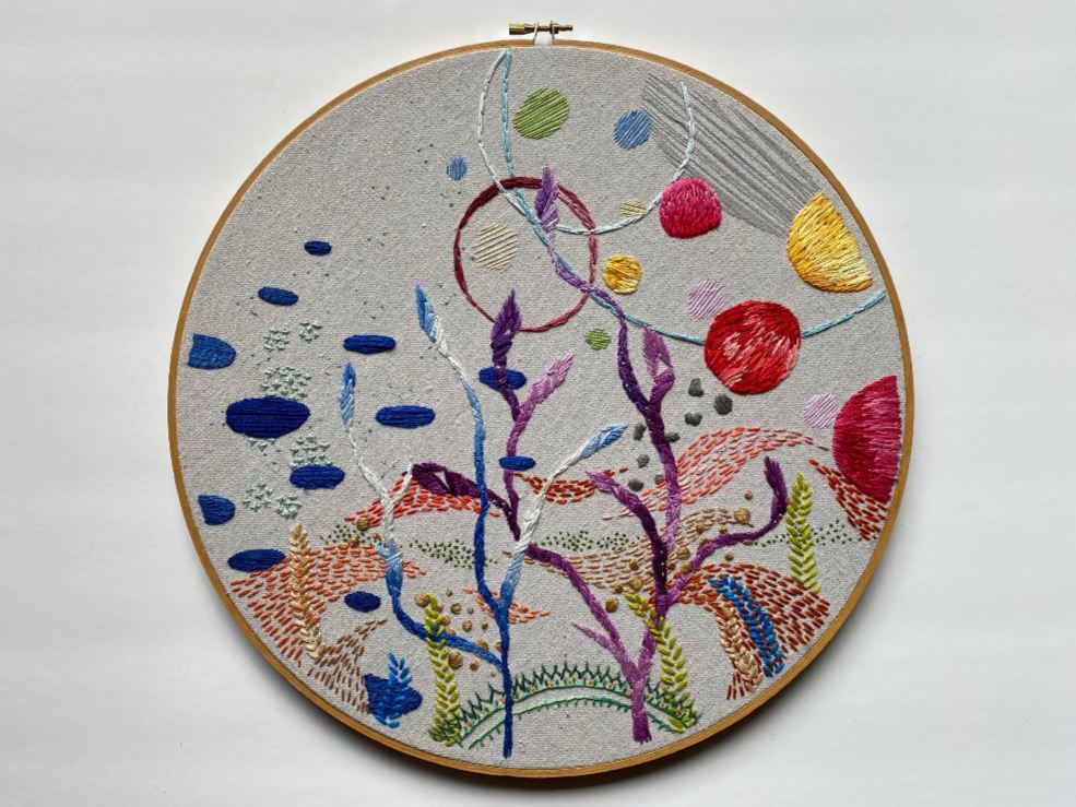 an embroidery hoop with gray fabric embroidered with a underwater seascape featuring all colors of the rainbow