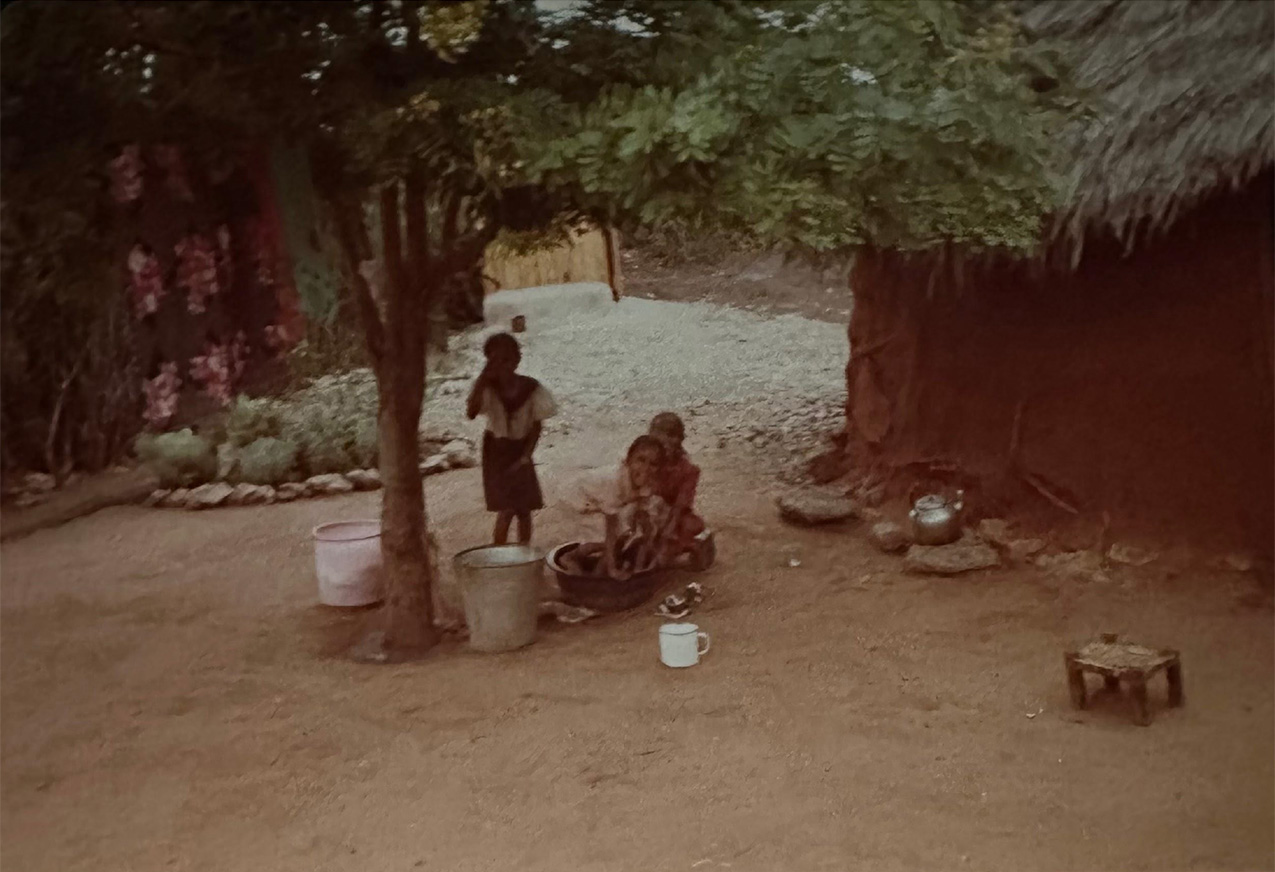 grainy photo of three children under a tree surrounded by bare dirt in Nairobi; one of the children squats in a large metal bowl used for bathing