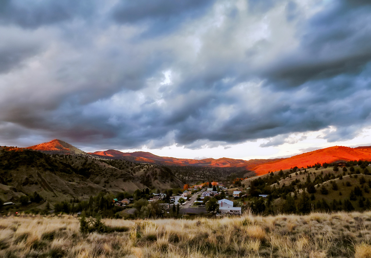 the city of Mitchell, Oregon nested between hills under a cloudy sky with distant peaks glowing orange from the sunset