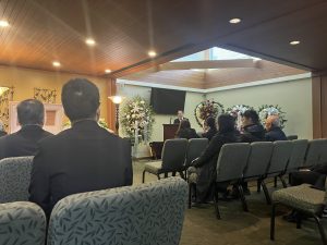 at a funeral, a man at a podium speaks to the seated mourners