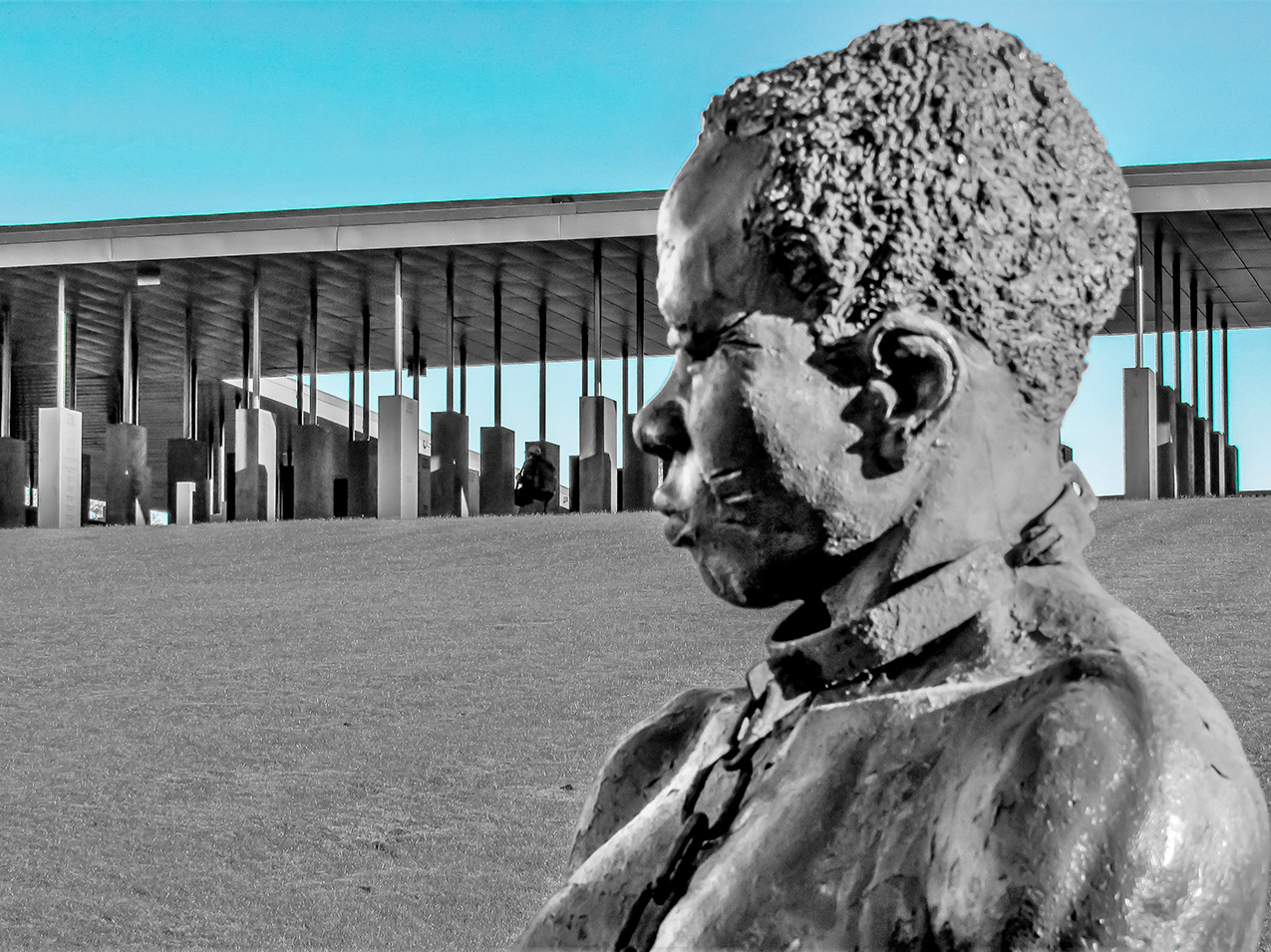 statue of an enslaved Black person in shackles in the foreground, with a memorial in the background. the photo is mostly black and white but the sky is turquoise blue.