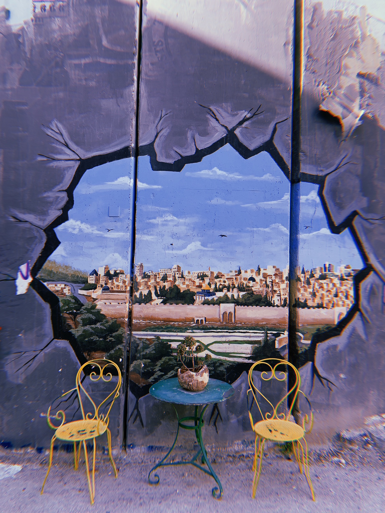 two bright yellow cafe chairs and table sit in front of a wall mural of Jerusalem as viewed through a hole in the wall