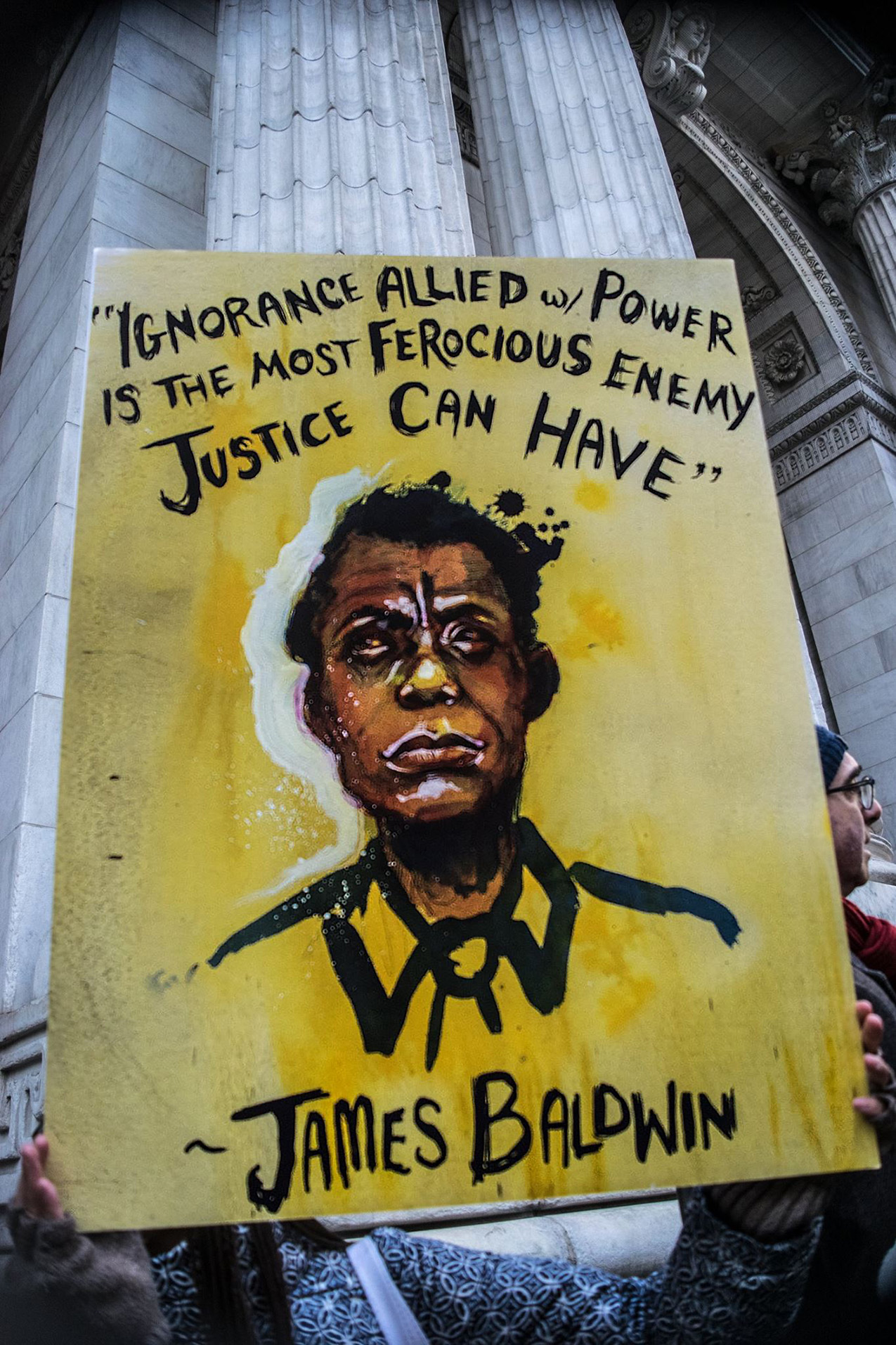 A protester holds up a yellow sign in front of a building with classical pillars. The hand-painted sign reads "Ignorance allied w/power is the most ferocious enemy justice can have –James Baldwin" with a painting of James Baldwin