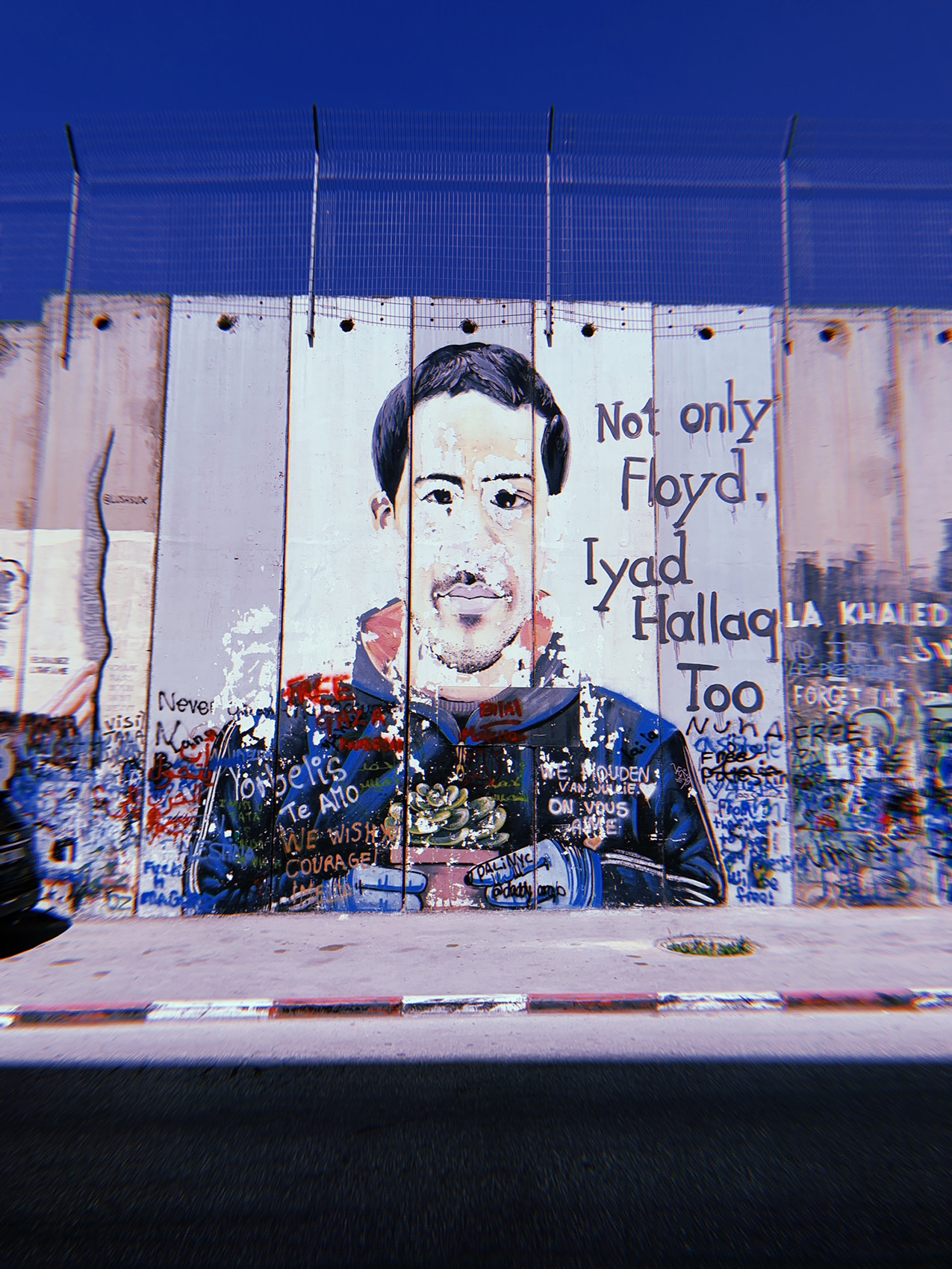 a wall with wire extending its height features a mural featuring a man's portrait with the text "Not only Floyd. Iyad Hallaq Too." The bottom portion of the wall and mural are covered in graffiti.