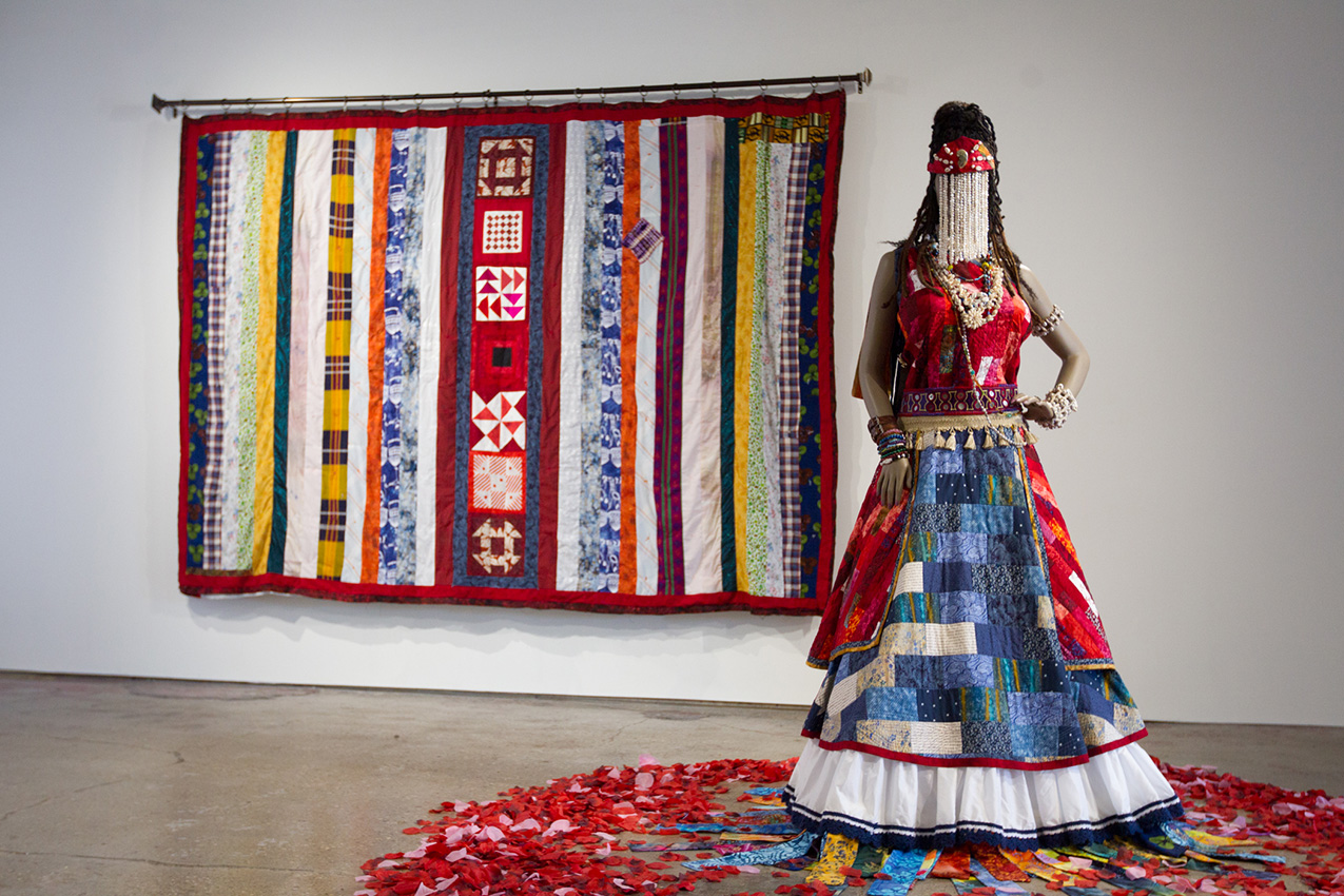 art installation featuring a mannequin wearing masquerade dress and a wall quilt, both primarily red with white, blue, and yellow accents