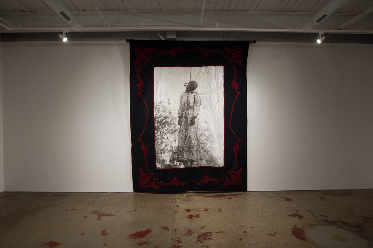 art installation of a quilt with a thick red and black border and a central black and white photo of a Black woman hanging from a noose