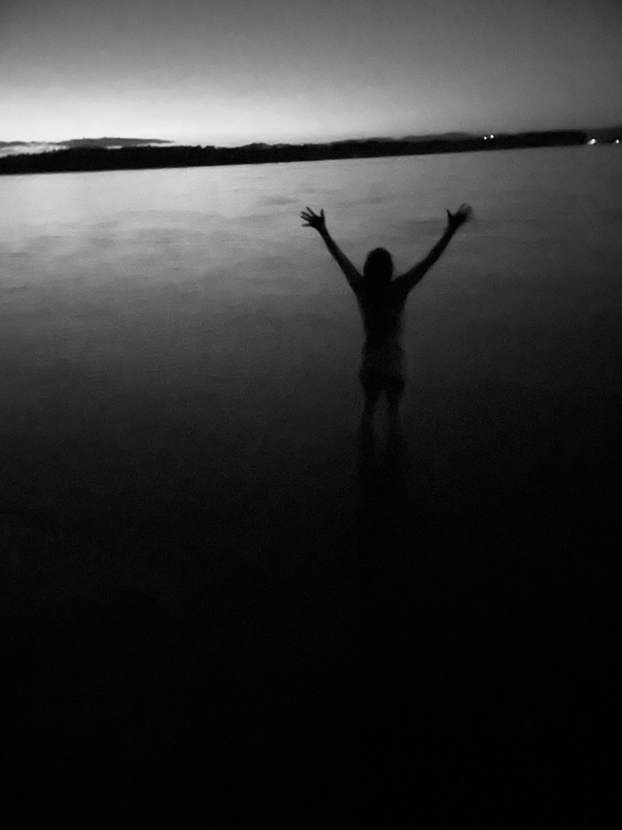 soft-focus, low-light black and white photo of a figure with its arms raised, silhouetted in front of a lake at night