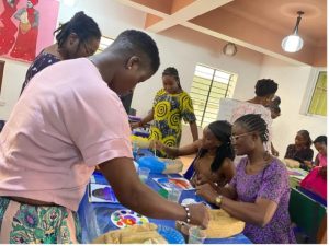 Black women, some standing and some sitting, pain wooden masks at a table covered with a blue tablecloth