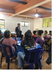 a man in a suit stands at the front of a room, while many women seated at tables listen to him teaching