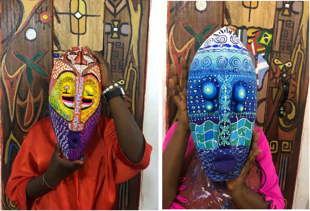 two Black women holding their brightly painted wooden masks in front of their faces