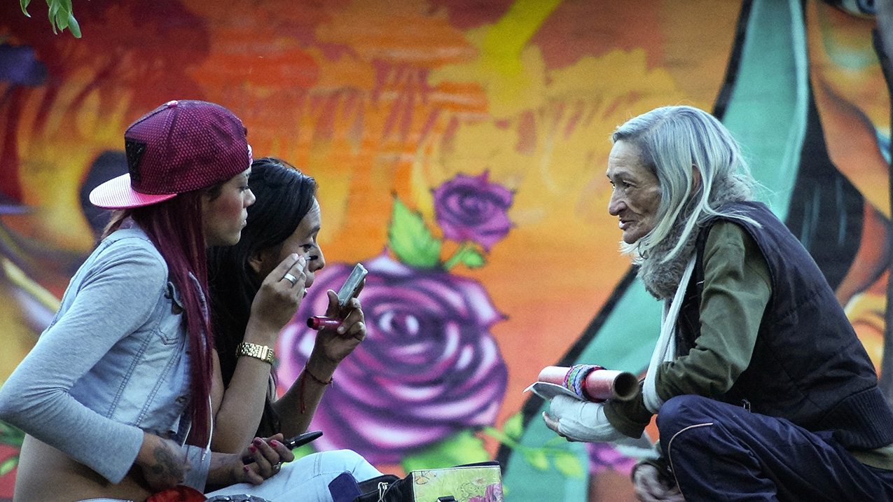 a gray-haired woman squats, facing two younger women, one of whom is applying mascara, in front a bright orange mural with a large purple rose