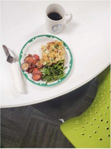 Plate with croissant casserole, rainbow carrots, and sauteed napini on a white table with a mug of coffee and lime green chair. 