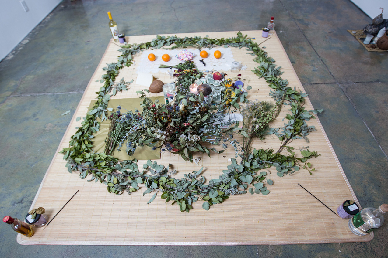 an altar of various offerings on a bamboo mat; the offerings include eucalyptus branches, fruit, bouquets of fresh and dried flowers; the corners of the mat are held down with bottles of liquor, jars, and incense sticks