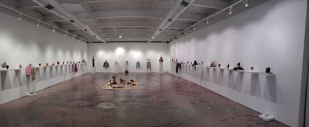 a concrete-floored gallery with white walls filled with simple while shelves, each holding a small figure and accessories; an altar is in the center of the floor