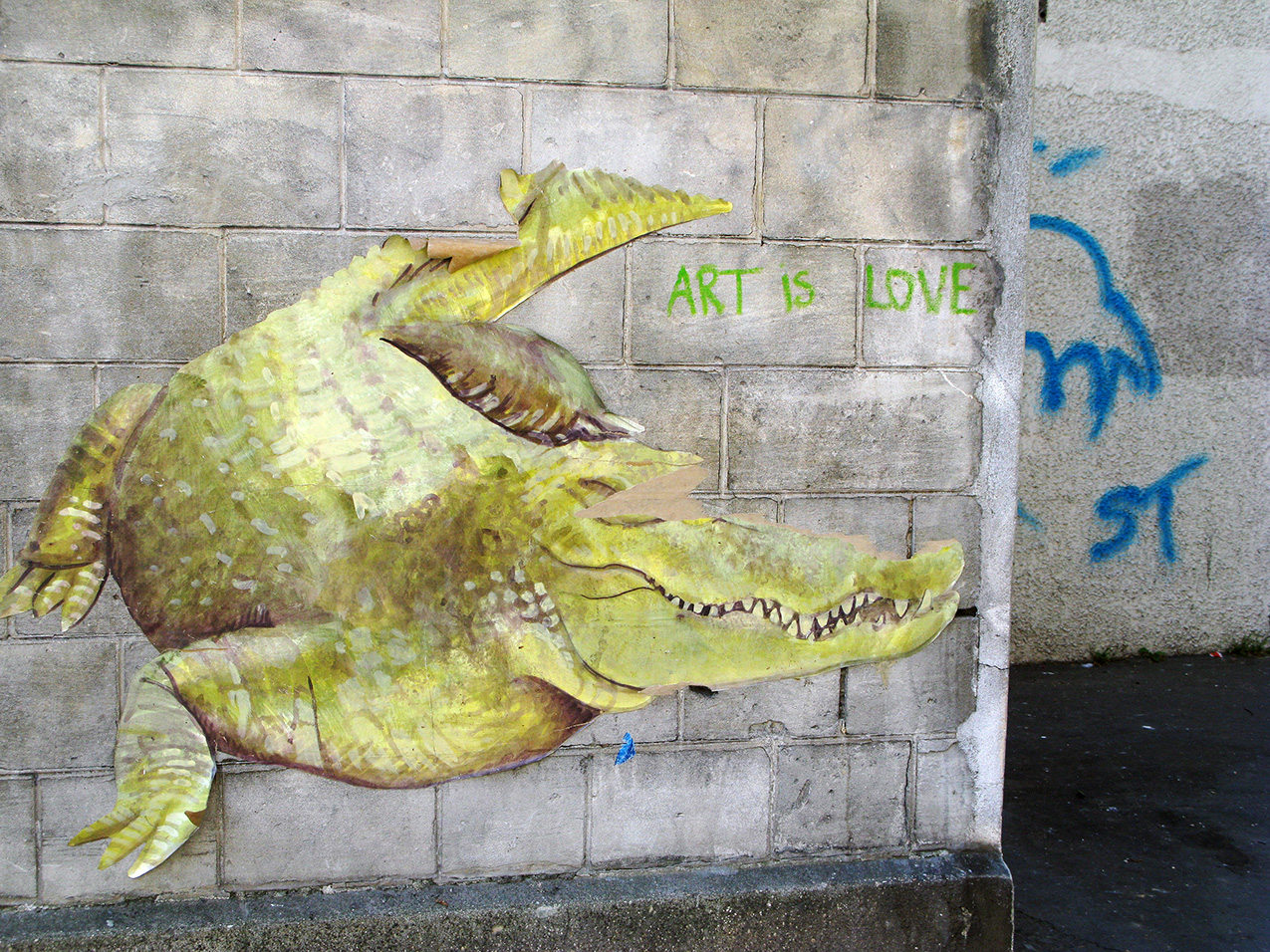 street art on a cement block wall: a paper applique yellow-green crocodile with peeling in the eye and tail areas; "ART IS LOVE" handwritten in green chalk next to it. a second wall, visible beyond the first, has the letters "ST" and an unrecognizable shape in blue spray paint.