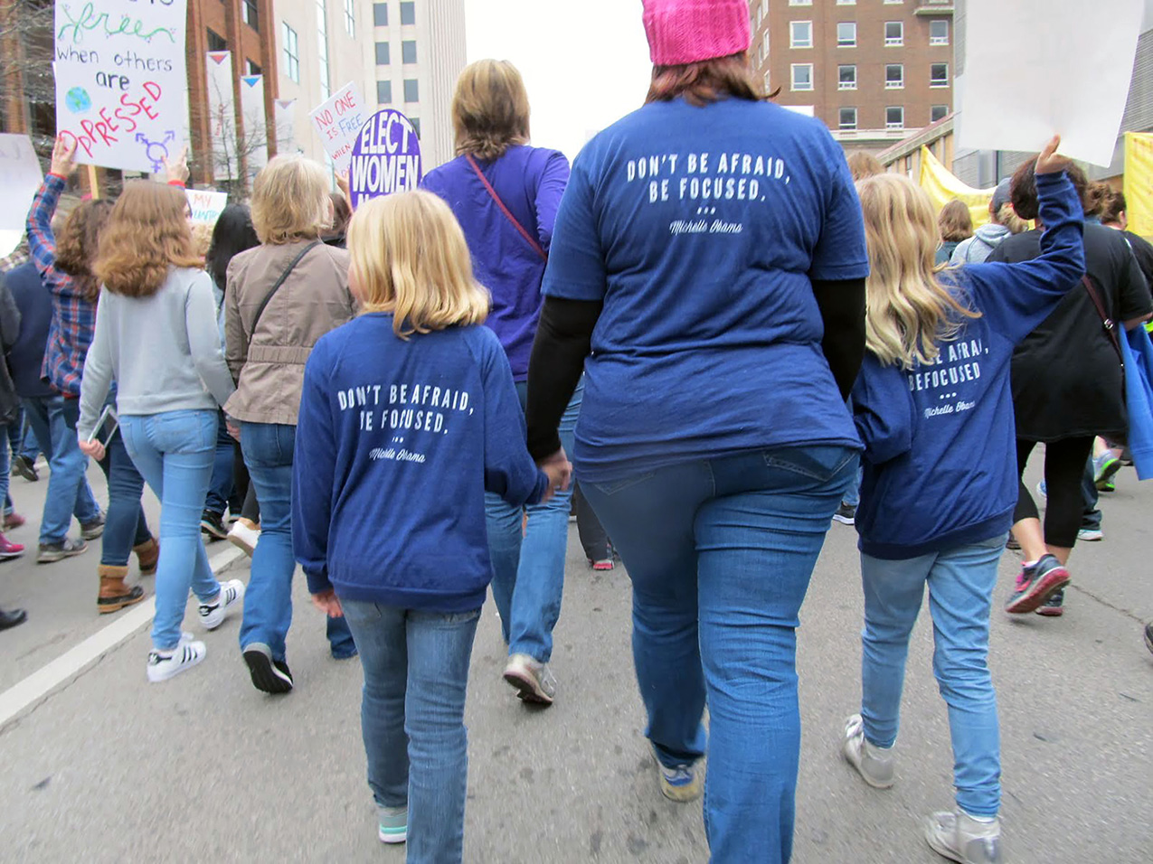 women marching on a city street holding signs. closest to the camera is a woman and her two young daughters who all wear blue shirts reading "DON'T BE AFRAID, BE FOCUSED. --Michelle Obama" on the backs.