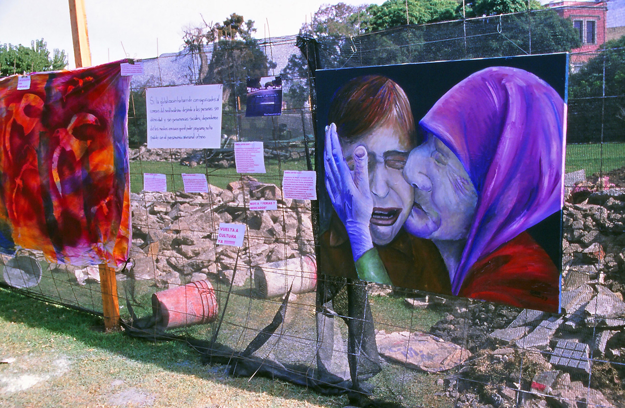 photo of artwork and signs hanging on a wire fence. on the left is a large piece of red batik fabric with seagulls silhouetted on it; on the right is a painting of an elderly woman in profile kissing the cheek of a younger crying woman. on the far side of the wire fence are piles of rubble, concrete bags, and buckets on a green lawn.