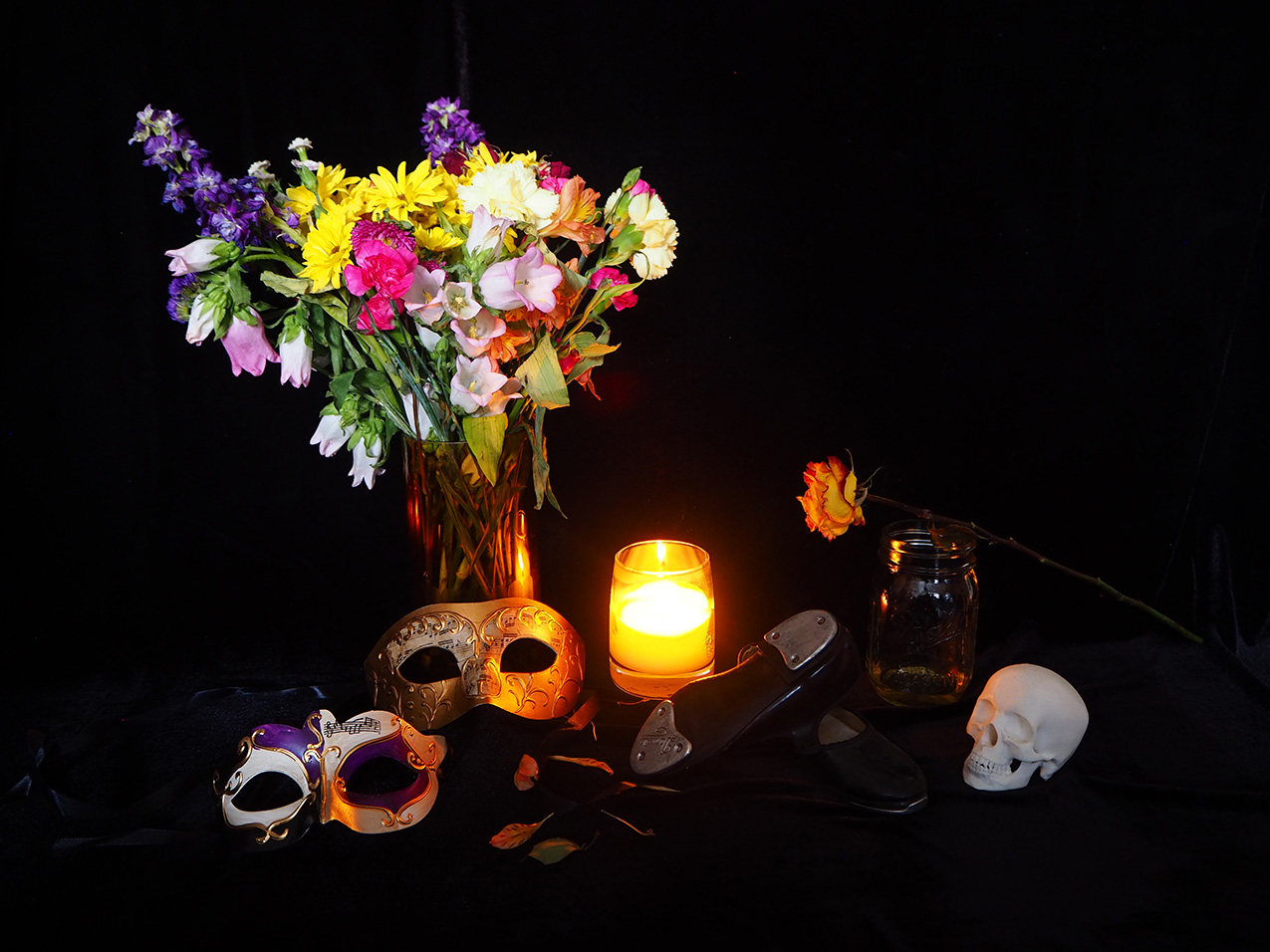 still life photograph on a black background: a bouquet of colorful flowers, two masquerade masks, a burning candle, tap shoes, a skull figurine and a single long-stemmed rose balanced on an empty jar