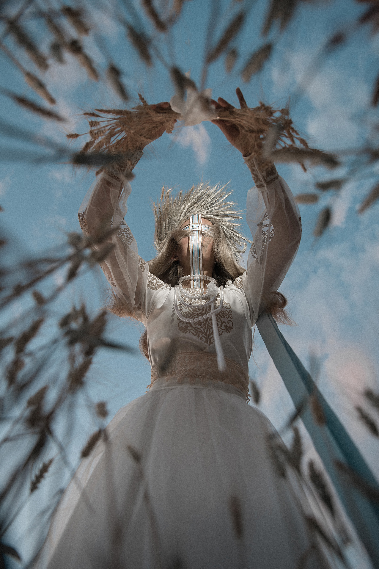 vertical photo looking up at a woman dressed in a traditional embroidered Ukrainian outfit, holding wheat in both outstretched hands. her arms form a circle framing her face, which is covered by ribbons in a cross shape. the background is a blue sky with light clouds; the foreground is shaped by strands of wheat that are out of focus.