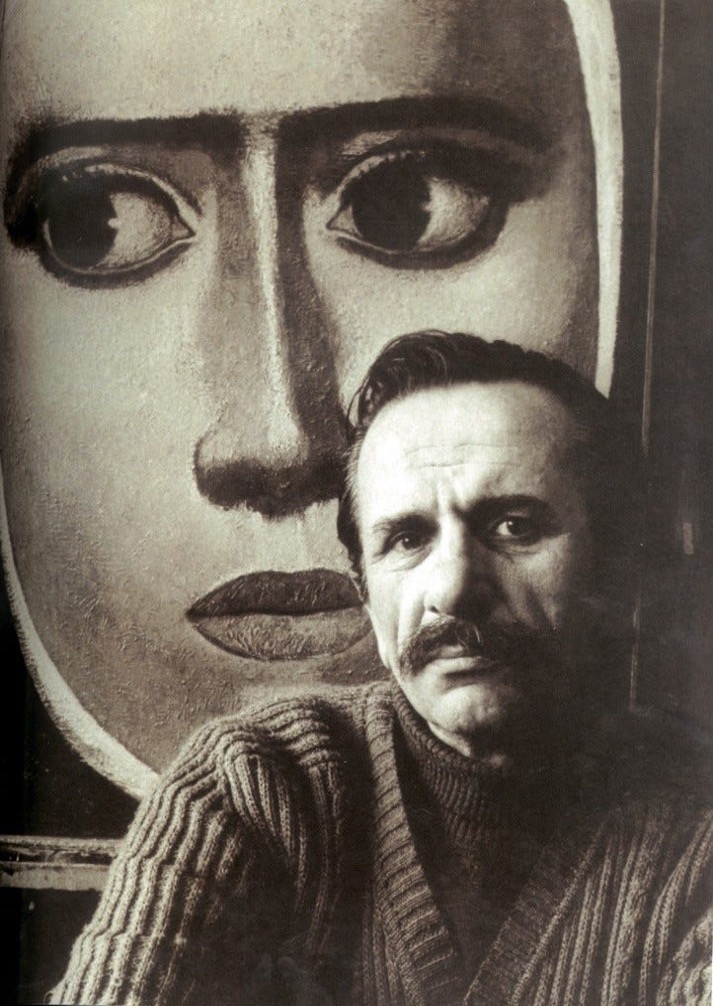 black and white photo of Nuri İyem, a man with a mustache and slicked hair wearing a ribbed sweater, in front of one of his paintings, a close-up of a woman's face, her large dark eyes looking to the left and brow slightly furrowed