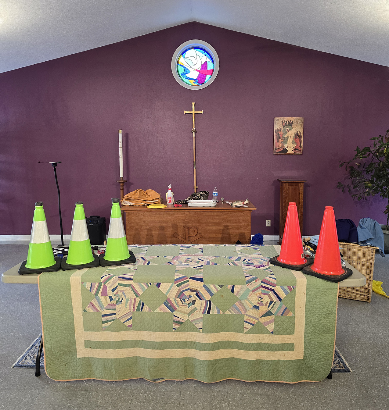 A minimal setting for a play: a pastel quilt draped over a table with construction cones on top of it. Located in an area that appears to typically serve as a church sanctuary: a purple wall with a peaked ceiling and circular stained glass window including objects such as a cross, candlestick, podium, and speaker.
