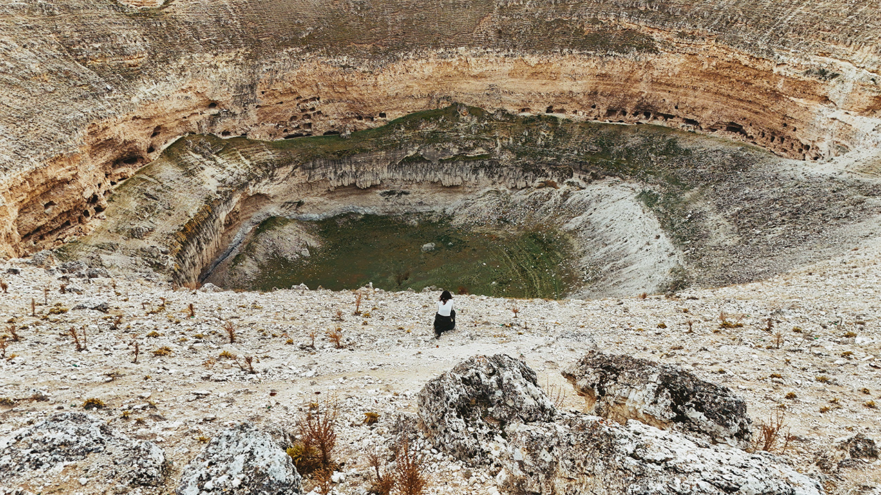 a woman sits near the edge of an obruk (giant sinkhole). the terrain is incredibly rocky, though the bottom of the sinkhole appears to have some green. some of the walls are cliff-like, others slope at a steep pitch.