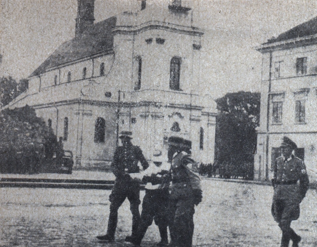 grainy black-and-white historical photo of a blindfolded prisoner being led by Nazi officers to his execution in front of bleachers of onlookers in a public square 
