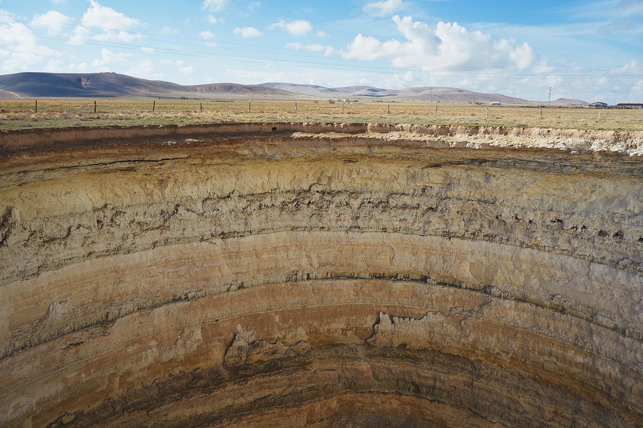 obruk (sinkhole) in a field. the top quarter of the photo is sky, distant rolling hills, and a field with fence posts. the bottom three-quarters of the image is striated layers of rock that form the walls of the sinkhole.