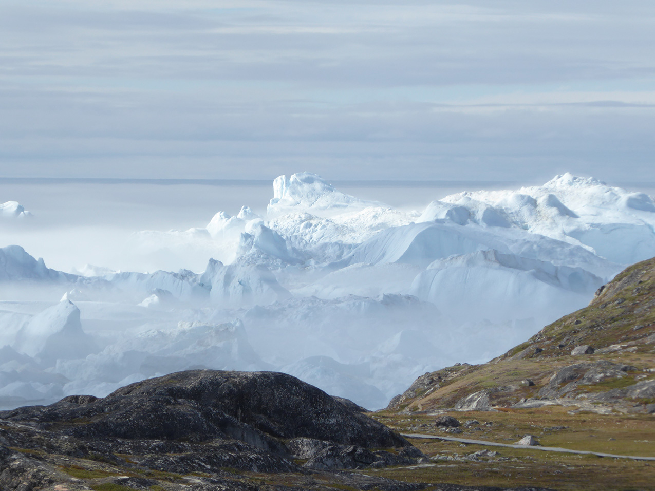 under a cloudy sky, a rugged glacier spans the width of the image. in the foreground, a path passes between rocky hills.