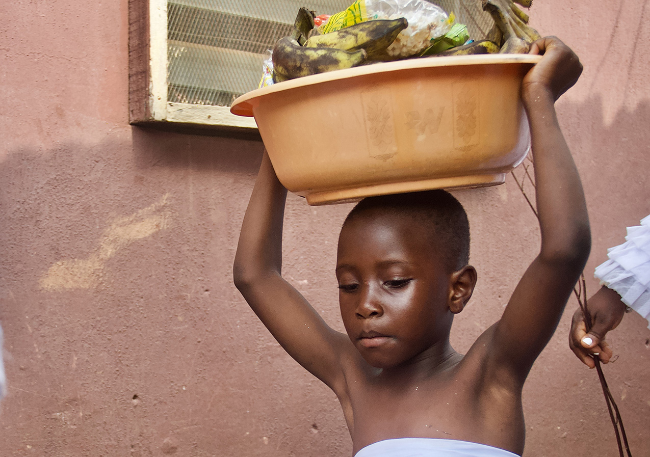 a young Black child with closely cropped hair concentrates on walking while holding a large basket filled with bananas and packaged food on their head