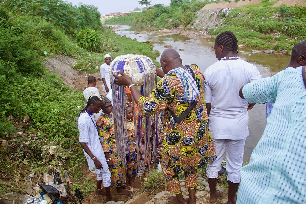 Black men and women, some dressed all in white and others dressed in colorful clothing, gather at a riverbank. one of the men in the foreground looks down to navigate the steep terrain while carrying a large drum or buoy shaped object covered in shells and long tassels