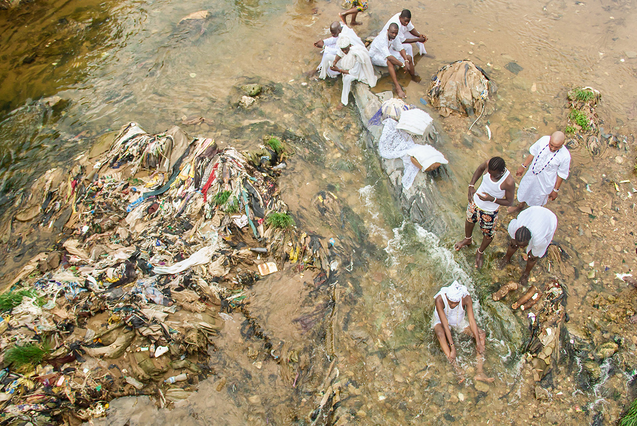 photo from above of eight Black men and women, dressed in white, standing or sitting in a shallow part of a river with offerings placed on a rock; on the left side of the frame, the water flows around what appears to be an pile of garbage