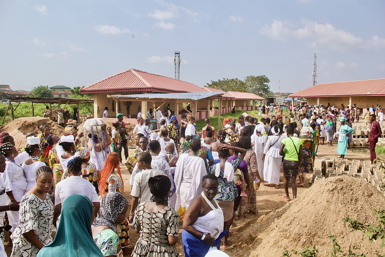 a crowd of African men and women walk away from the viewer toward a large one-story building with an adobe-colored roof; more people mingle near the building, making one hundred or more people in total