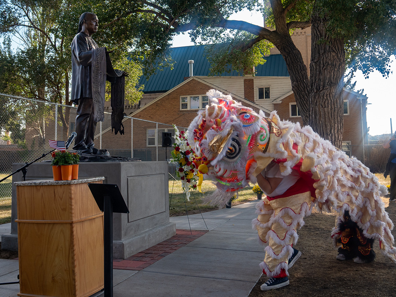 Two people in a lion dancer costume in front of the Requiem memorial statue, a bronze status of a Chinese man holding a draped flag. There is a church and tall fence in the background, and a podium in the foreground.