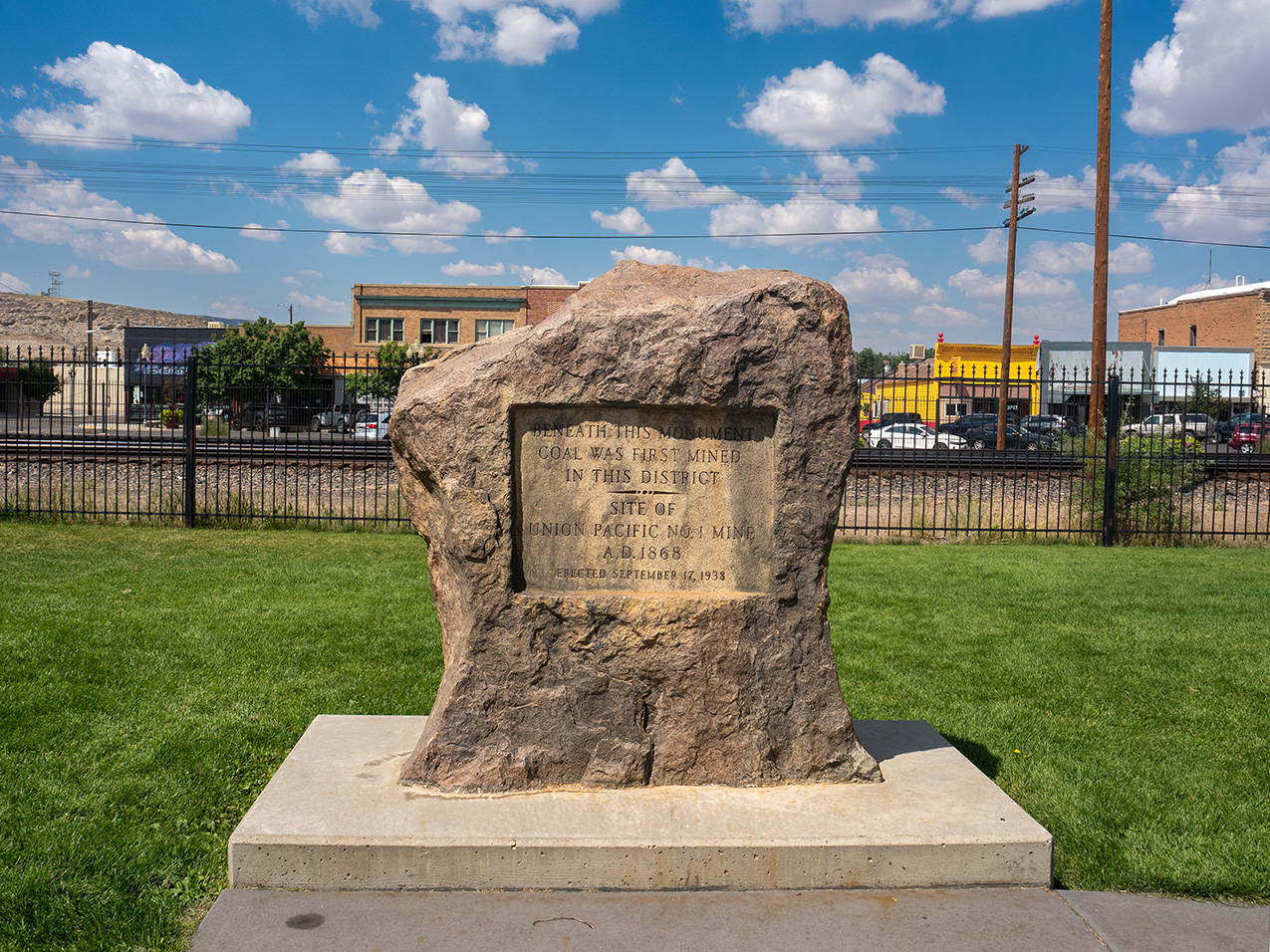 a large rock monument atop a concrete slab with a fence, railroad, and town in the background. the monument reads "Beneath this monument coal was first mined in this district. Site of Union Pacific No.1 Mine A.D.1868, erected September 17, 1938"