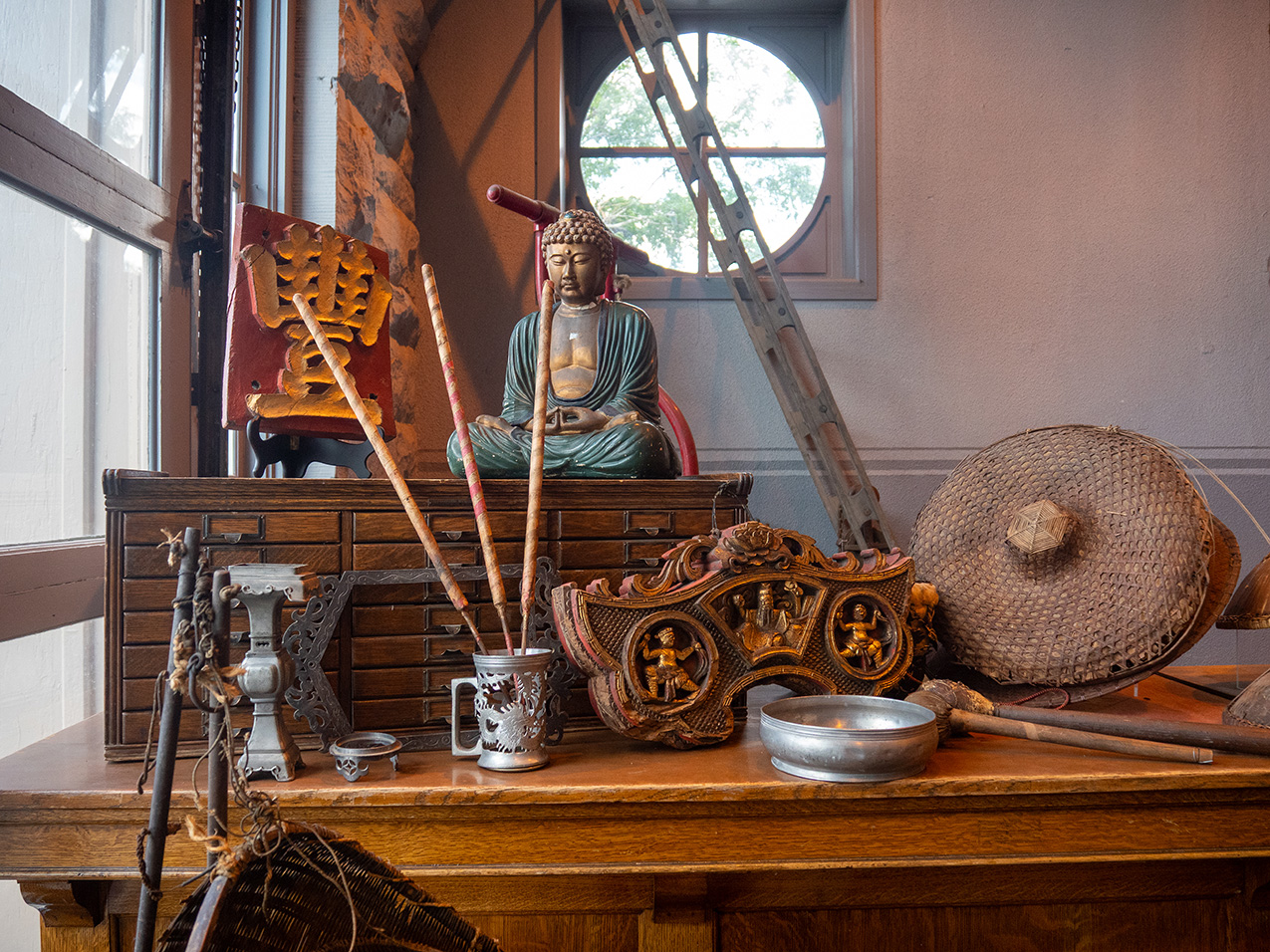a tabletop display of Chinese artifacts including a large Hanzi character made of wood, a buddha statue, a drum and mallets, decorative metal containers and carved wooden items.