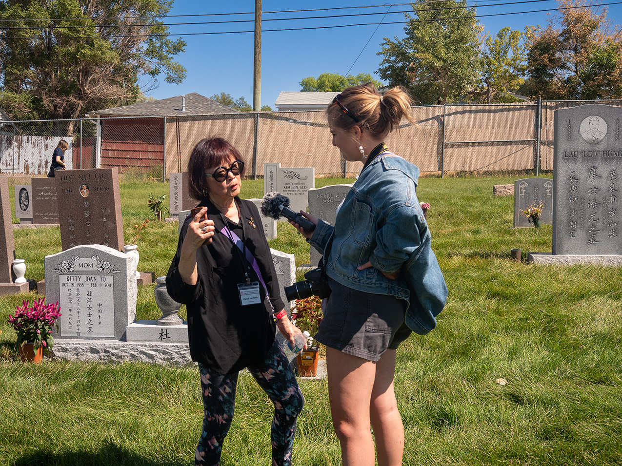 in a cemetery with Chinese characters carved on the headstones, a young blonde woman wearing a jean jacket and shorts holds out a microphone to a shorter Asian woman who is wearing floral tights, a black top, and a nametag. 